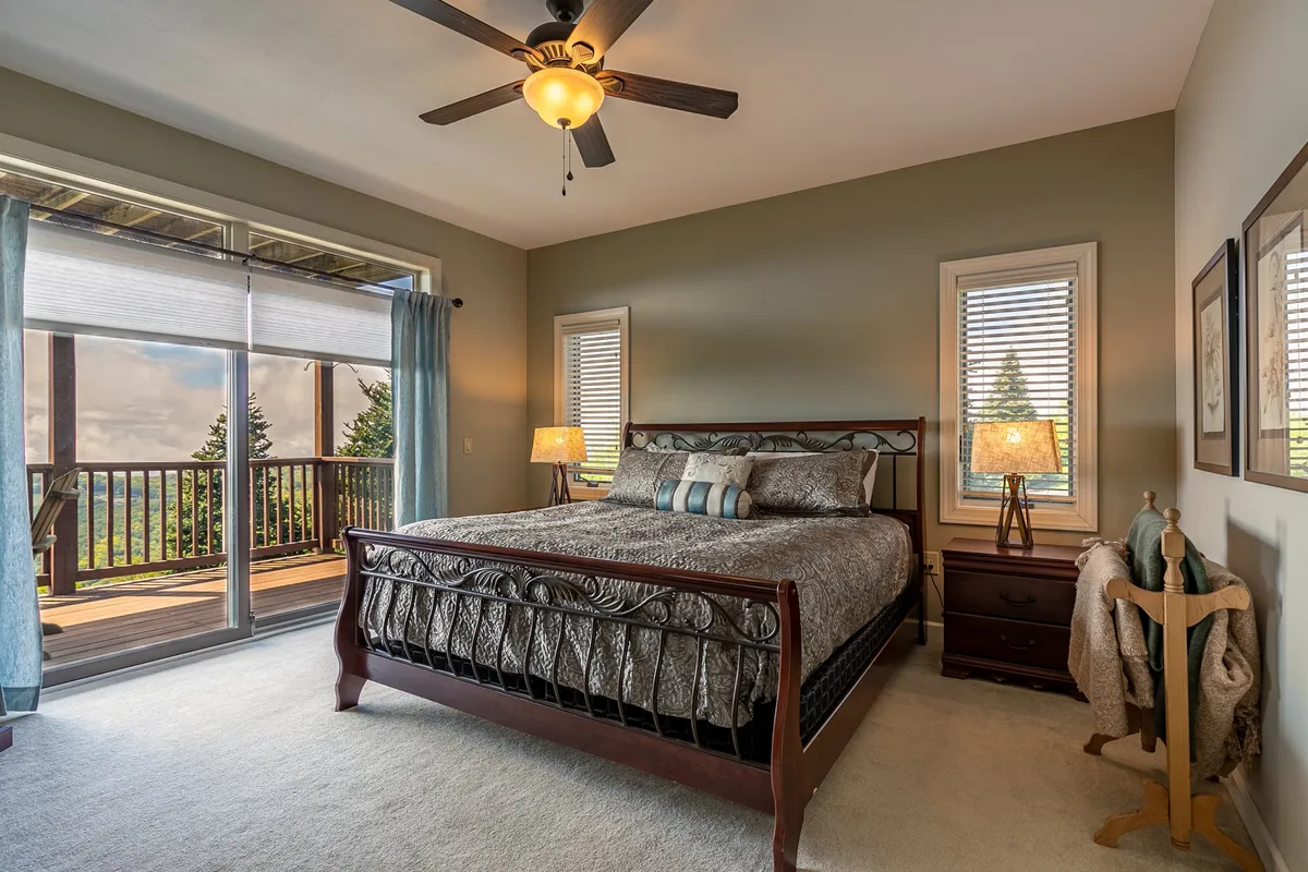 A tranquil bedroom featuring a neatly made bed with decorative pillows, large windows with curtains that invite natural light, and a nightstand with a glowing lamp. The expansive windows provide views outside, hinting at mountain vistas and cozy nights in.