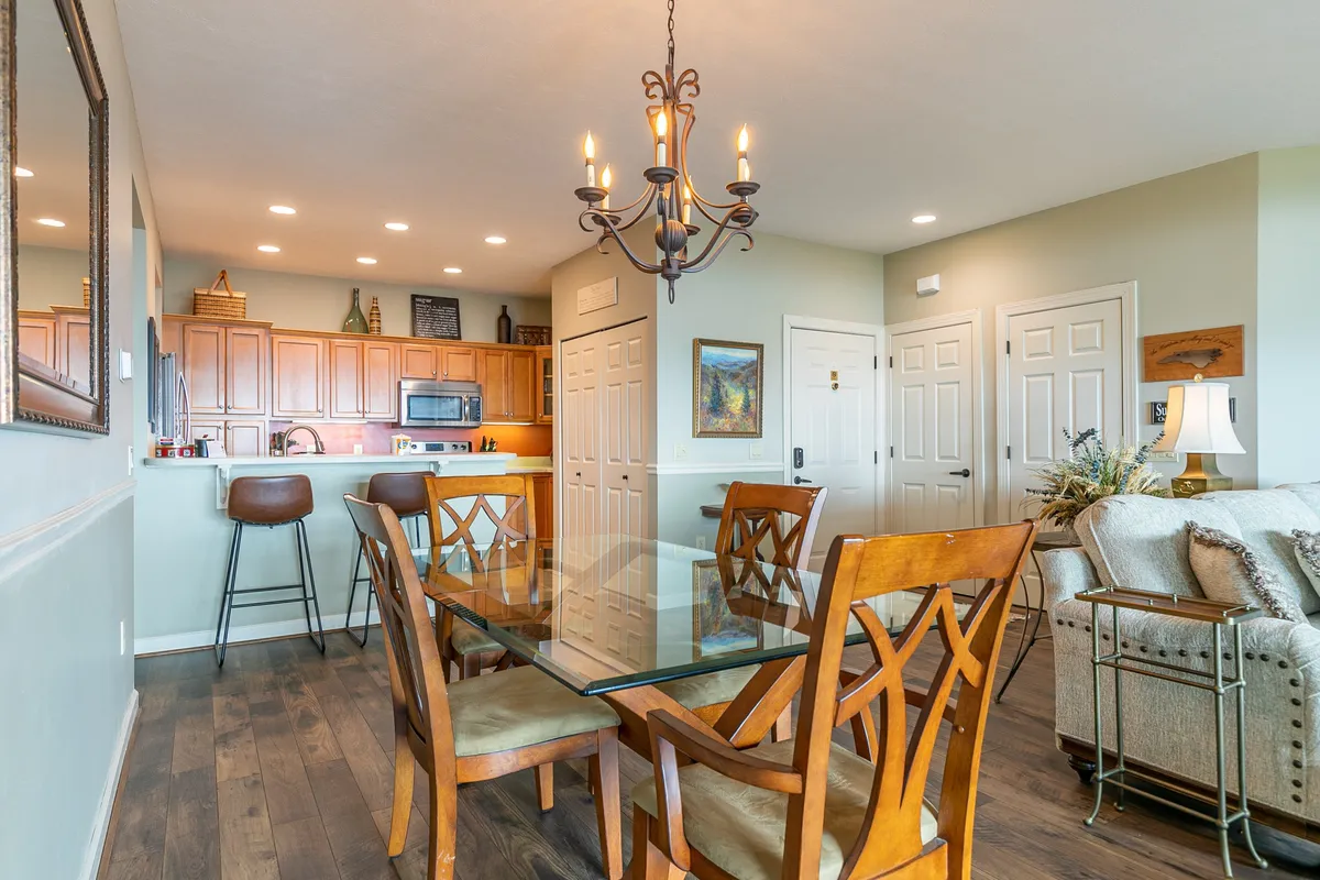 A bright dining area with a glass dining table, wooden chairs, a chandelier overhead, and a kitchenette in the background. The space feels open and is ideal for group meals or celebrations.