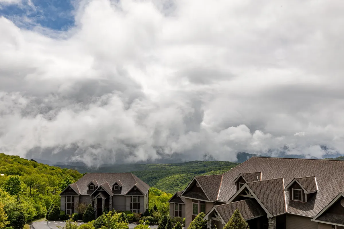 A tranquil exterior scene showcasing the architectural details of the SugarJax homes set against a breathtaking mountainous landscape, with soft clouds overhead and hints of autumn foliage in the surrounding hills. The foreground features a welcoming porch area ideal for relaxed outdoor gatherings and taking in sunrise or stargazing views.