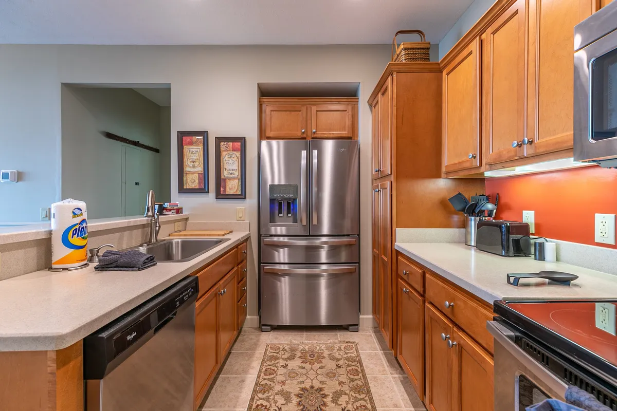 This image highlights the modern kitchen, featuring a stainless steel refrigerator, wooden cabinets, granite countertops with a sink, and a decorative rug. The space feels warm and welcoming, suggesting a perfect setting for cooking and entertaining.