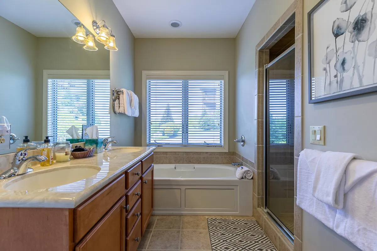 A spacious and serene bathroom with a double sink vanity, a soaking bathtub, a separate tiled shower area, and a window with blinds. The layout and finishes evoke a spa-like feel, perfect for unwinding after a day of adventure.