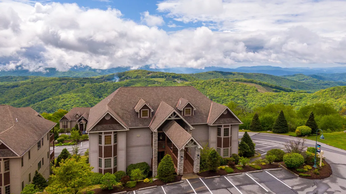 The image features the exterior of a well-maintained condo building set against a stunning mountainous backdrop. Lush, manicured landscaping surrounds the building, and architectural details hint at upscale comfort. The tranquil scene highlights the natural beauty of Sugar Mountain, inviting guests to relax, explore hiking trails, and savor breathtaking sunsets.