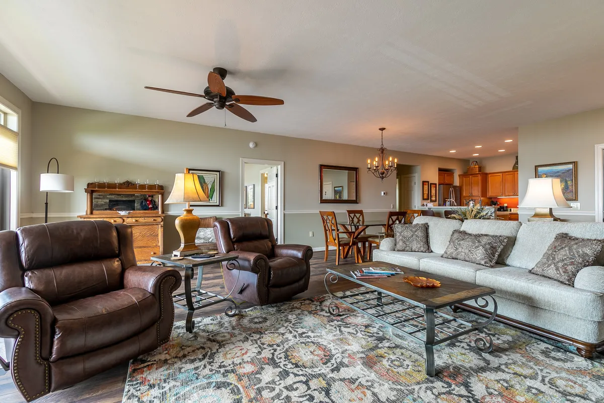 This wide-view image captures the transition from the kitchen/dining area to the adjacent living space, with recliner chairs, a coffee table, and a dining area visible. The arrangement suggests a seamless flow between cooking, dining, and relaxing, while large windows offer an inviting glimpse outside.