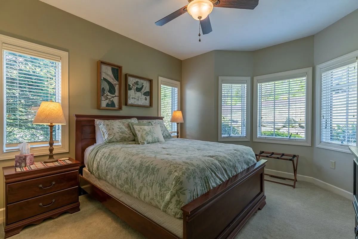 A serene bedroom filled with natural light, featuring floral bedding, a bedside lamp, and windows overlooking nature. The peaceful mood suggests restful nights and gentle mornings.