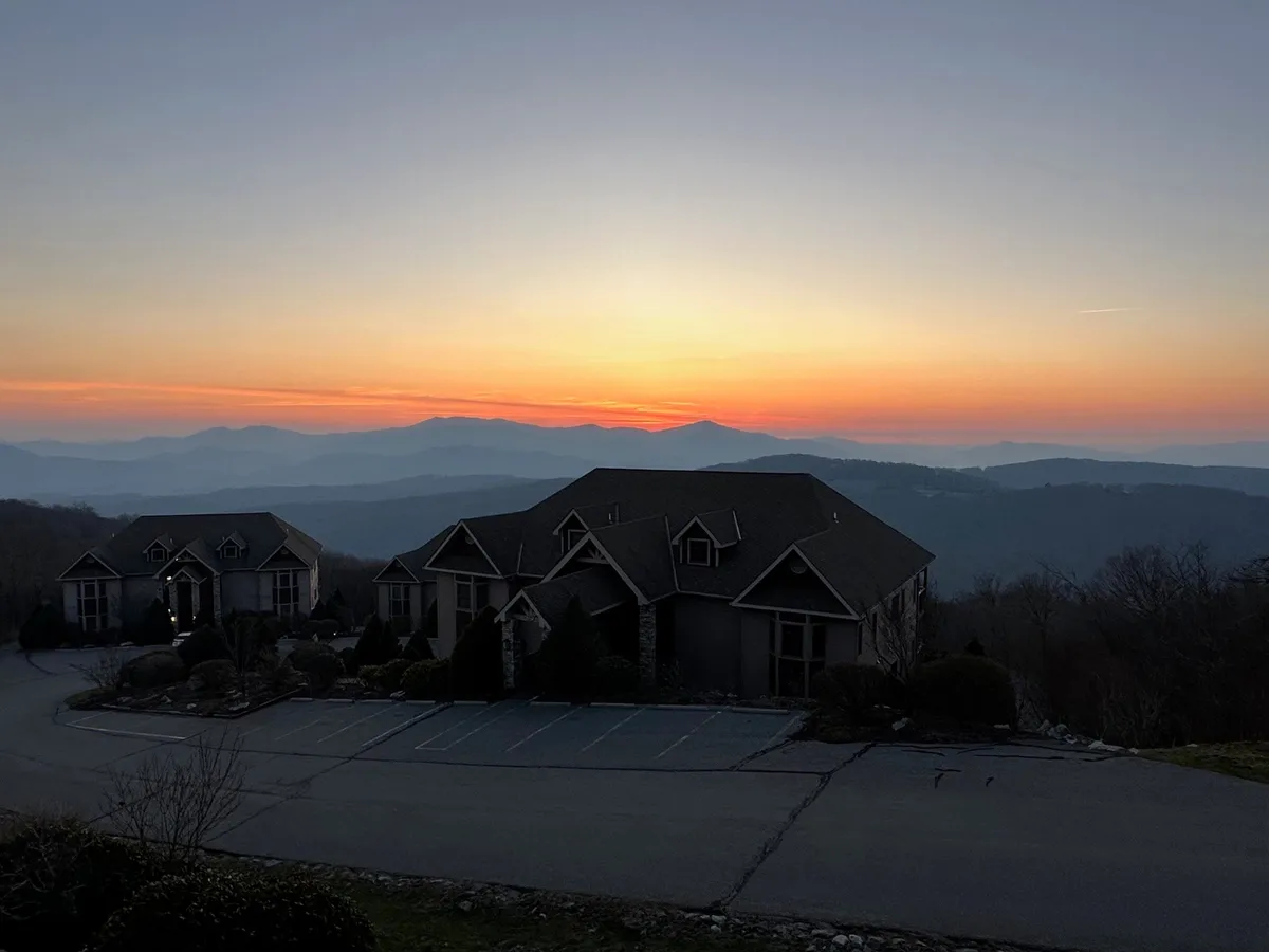 An exterior perspective showcasing a row of homes with porches oriented toward a sweeping mountain vista, accented by a vibrant sunset sky. The scene emphasizes relaxation and connection to the landscape, evoking feelings of tranquility and shared experience.